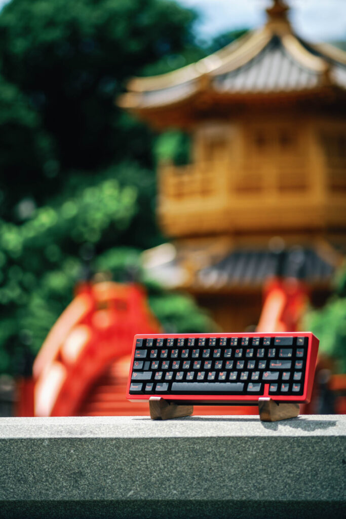 Red-framed 60 percent mechanical keyboard with black keycaps on a wooden stand before a blurred golden pagoda