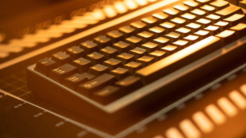 Compact dark mechanical keyboard with illuminated keycaps on a grid-lined surface under warm golden lighting