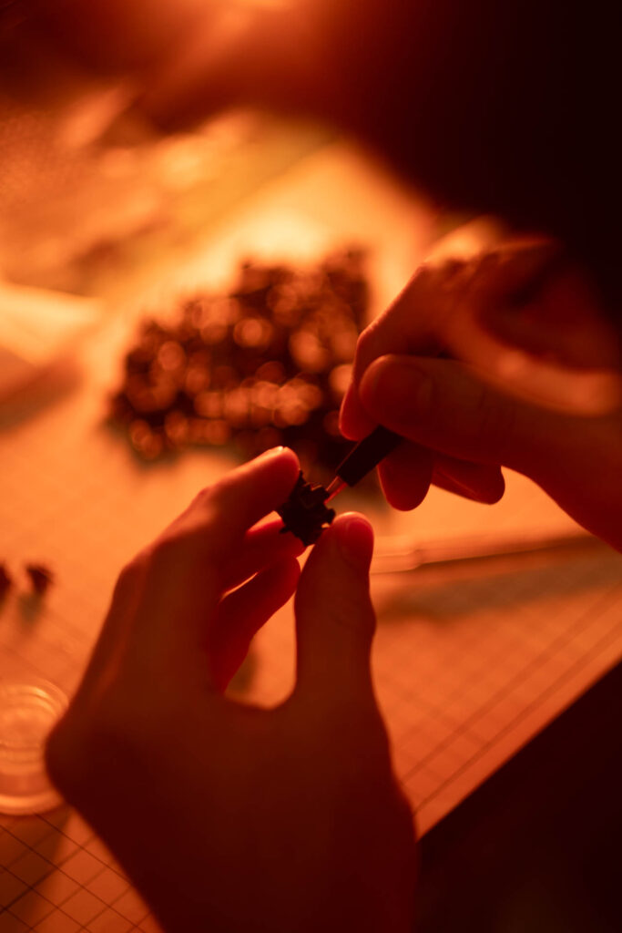 Hands applying lubricant to a black switch on a white grid-lined keyboard under warm orange lighting
